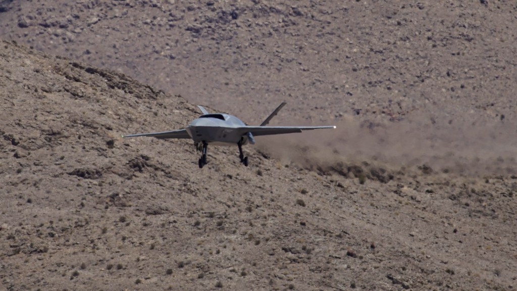 A gray uncrewed jet with its landing gear down descends past a rocky desert hillside.