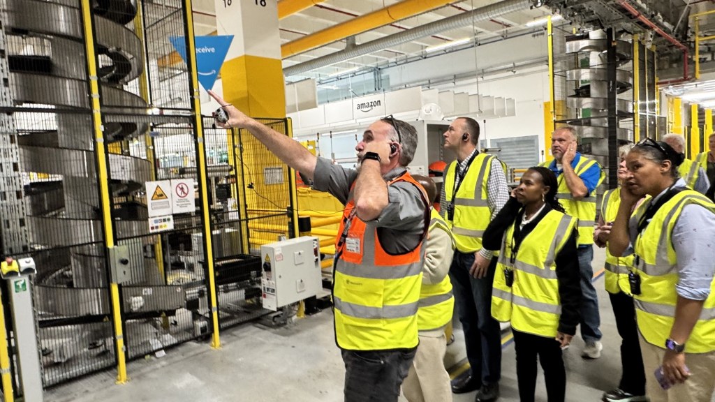 A man in a high-visibility vest points toward automated conveyor equipment while visitors in yellow vests look on inside an Amazon warehouse.