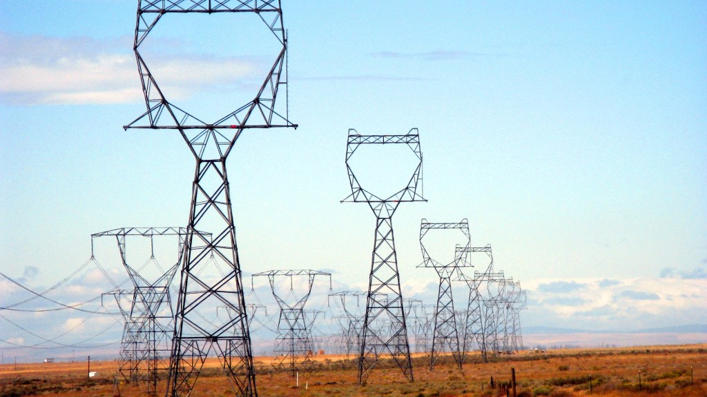 A row of steel high-voltage transmission towers crossing dry grassland under a blue sky.