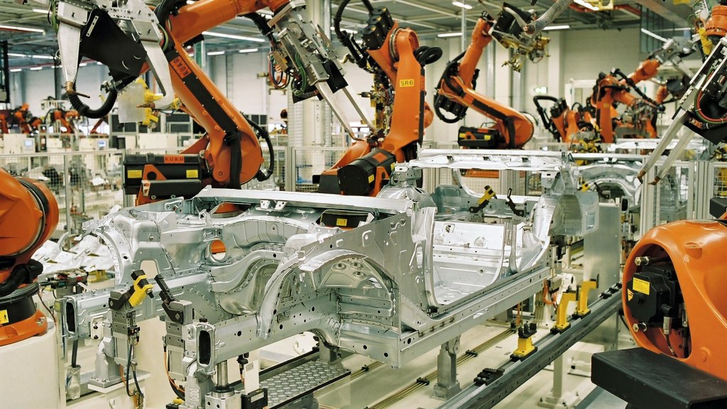 Rows of orange industrial robot arms welding car body frames on an automotive assembly line.