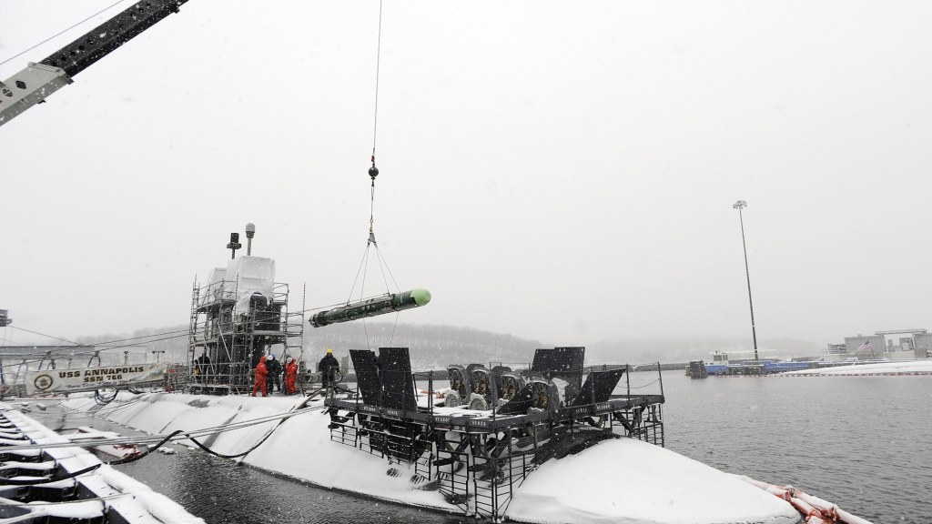 Sailors in cold-weather gear guide a long Mk 48 torpedo suspended from a crane beside a submarine during snowfall.