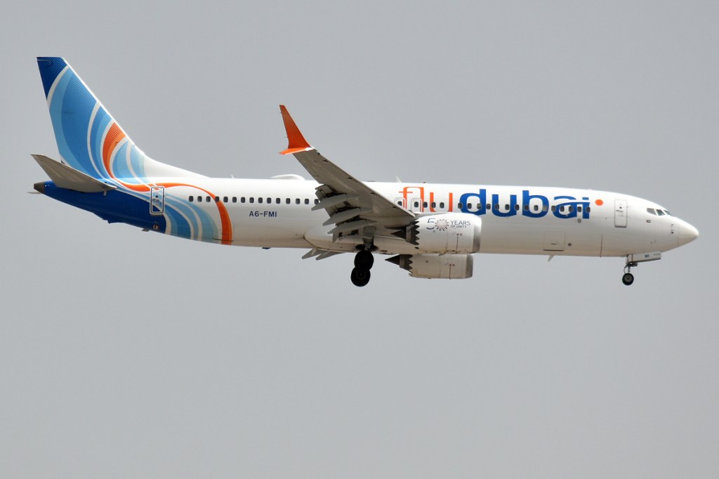 A white flydubai Boeing 737 MAX with blue and orange tail livery flies with its landing gear down against a gray sky.