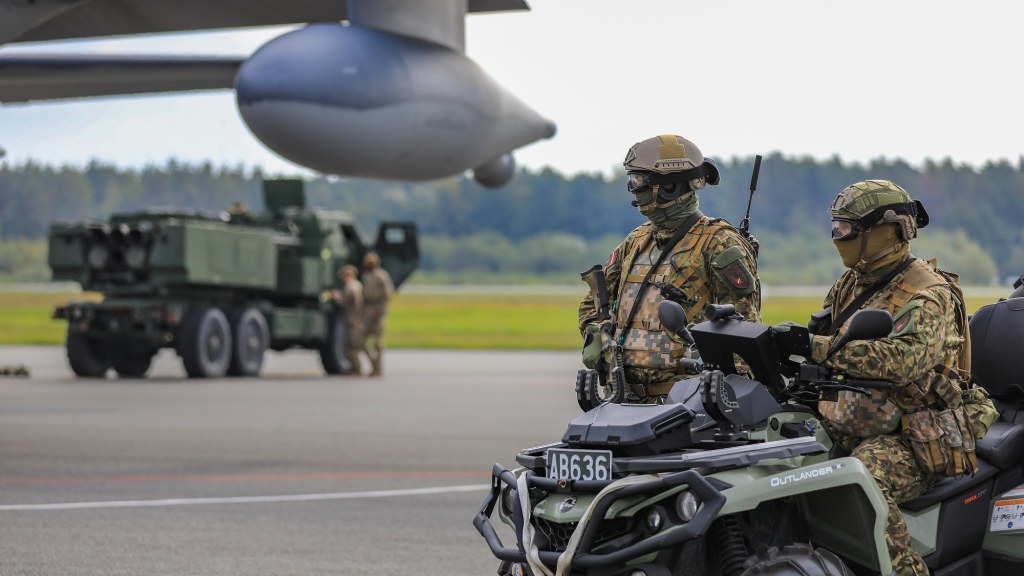 Two Latvian soldiers on an all-terrain vehicle in the foreground with a HIMARS rocket launcher truck and a transport plane behind them.