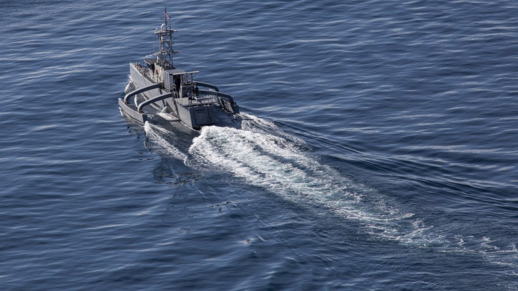 A long, narrow gray uncrewed trimaran vessel cuts through the ocean with no crew visible on deck.