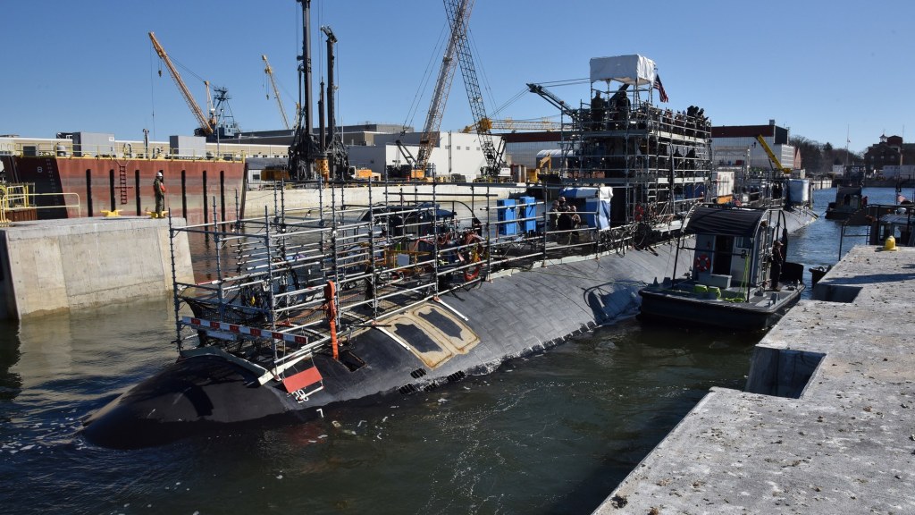 A submarine moves through a narrow channel into a flooded dry dock basin, with shipyard cranes and buildings around it.