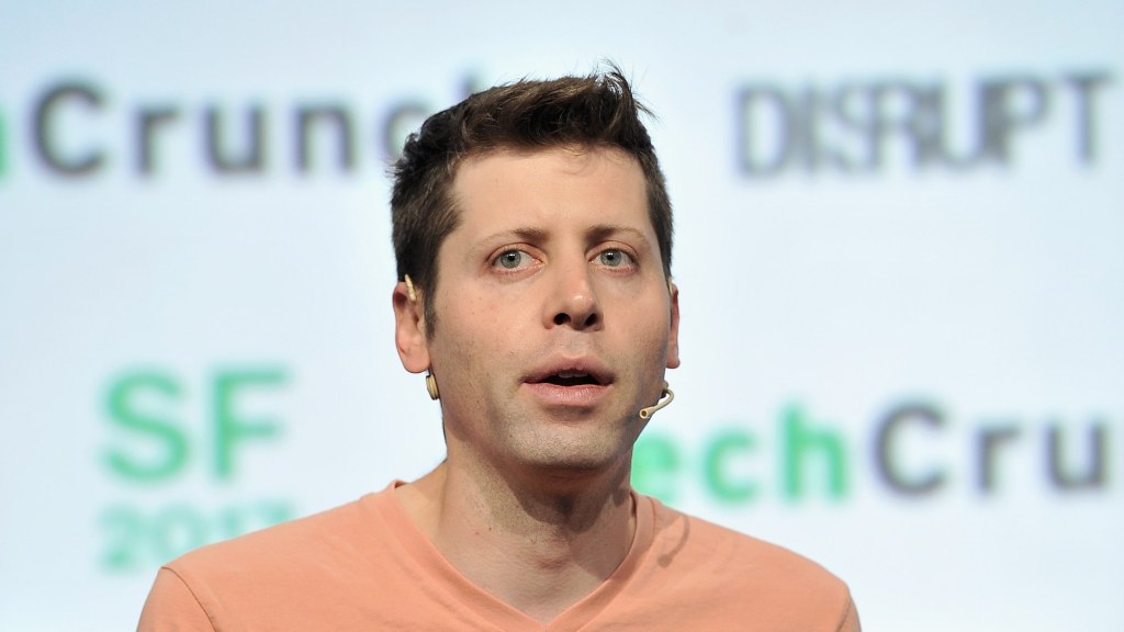 Sam Altman, wearing a headset microphone and an orange T-shirt, speaks on a stage in front of a blurred event backdrop.