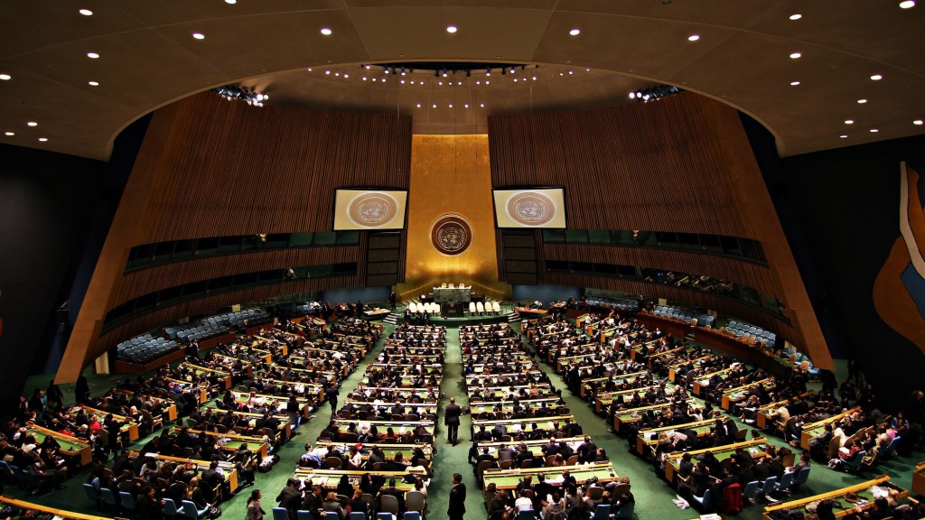The empty U.N. General Assembly Hall with rows of delegate desks facing the podium and U.N. emblem.