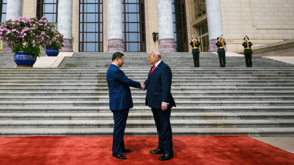 Donald Trump and Xi Jinping shake hands on a red carpet at the foot of the steps of the Great Hall of the People, with a military honor guard behind them.