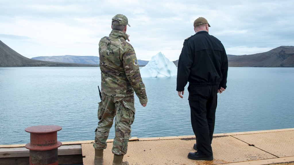 Two uniformed officers stand on a pier at Pituffik Space Base looking toward an iceberg in a fjord ringed by hills.