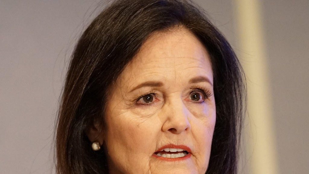Close-up of Judy Shelton, a woman with shoulder-length dark hair and pearl earrings, speaking at a conference.