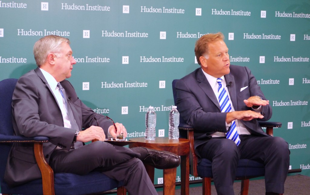 Mike Rogers, in a dark suit and blue striped tie, gestures while seated on a stage in front of a Hudson Institute backdrop, next to another man in an armchair.