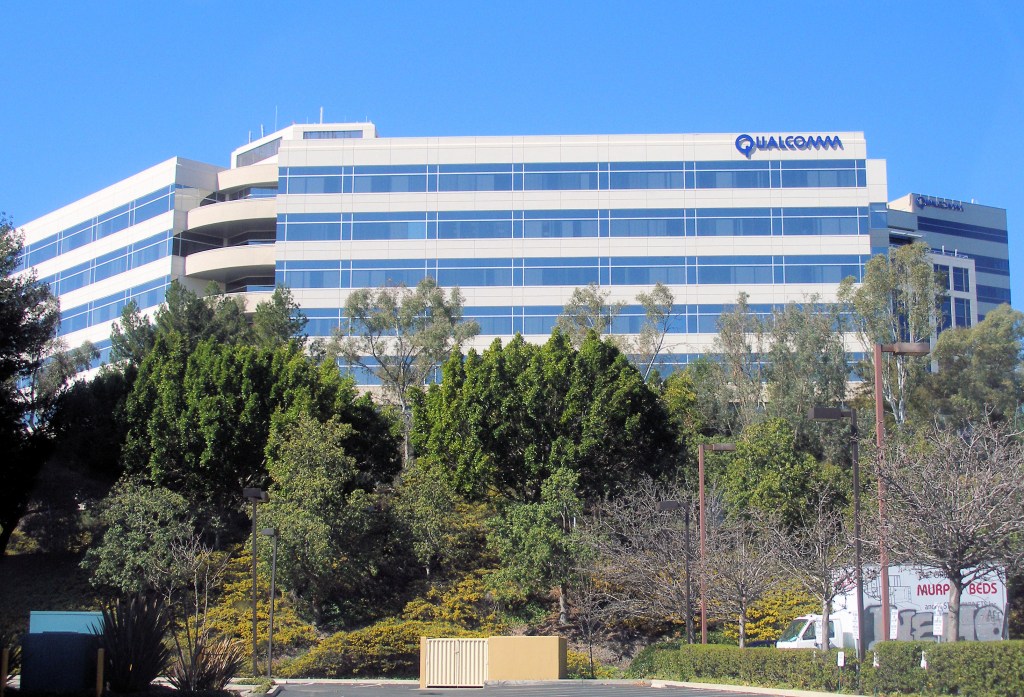 A white and blue-glass Qualcomm office building with the company logo near the roofline, behind a row of trees.