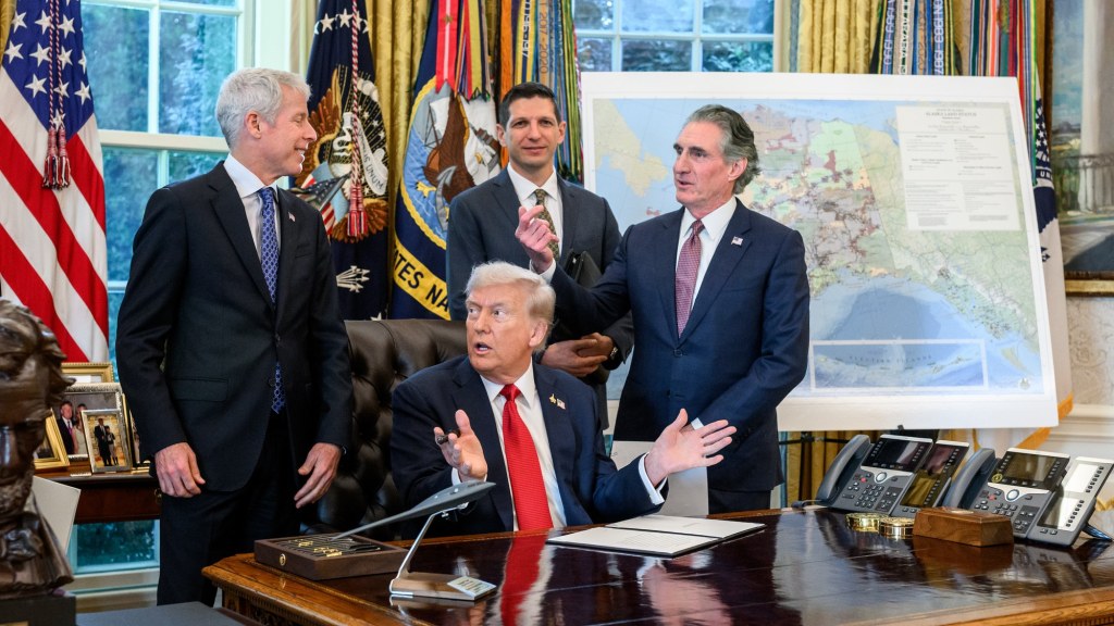 President Trump, seated at the Resolute Desk in the Oval Office, gestures while officials stand behind him beside a map on an easel.