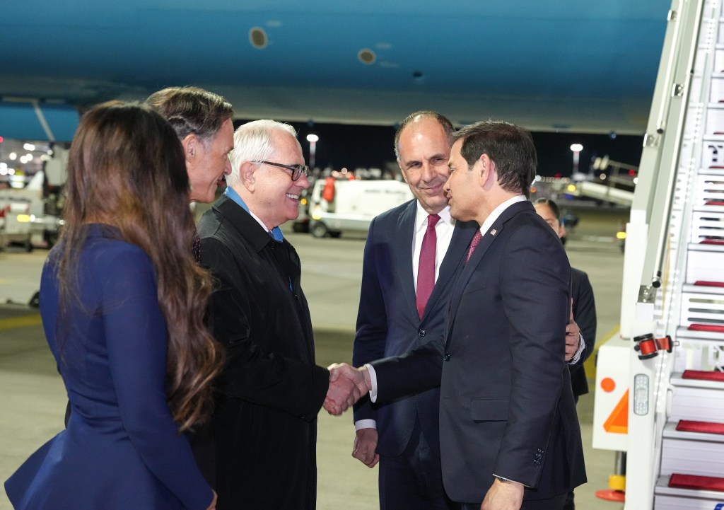 Secretary of State Marco Rubio, in a dark suit, shakes hands with a greeting party at the foot of an aircraft stairway on an airport tarmac at night.